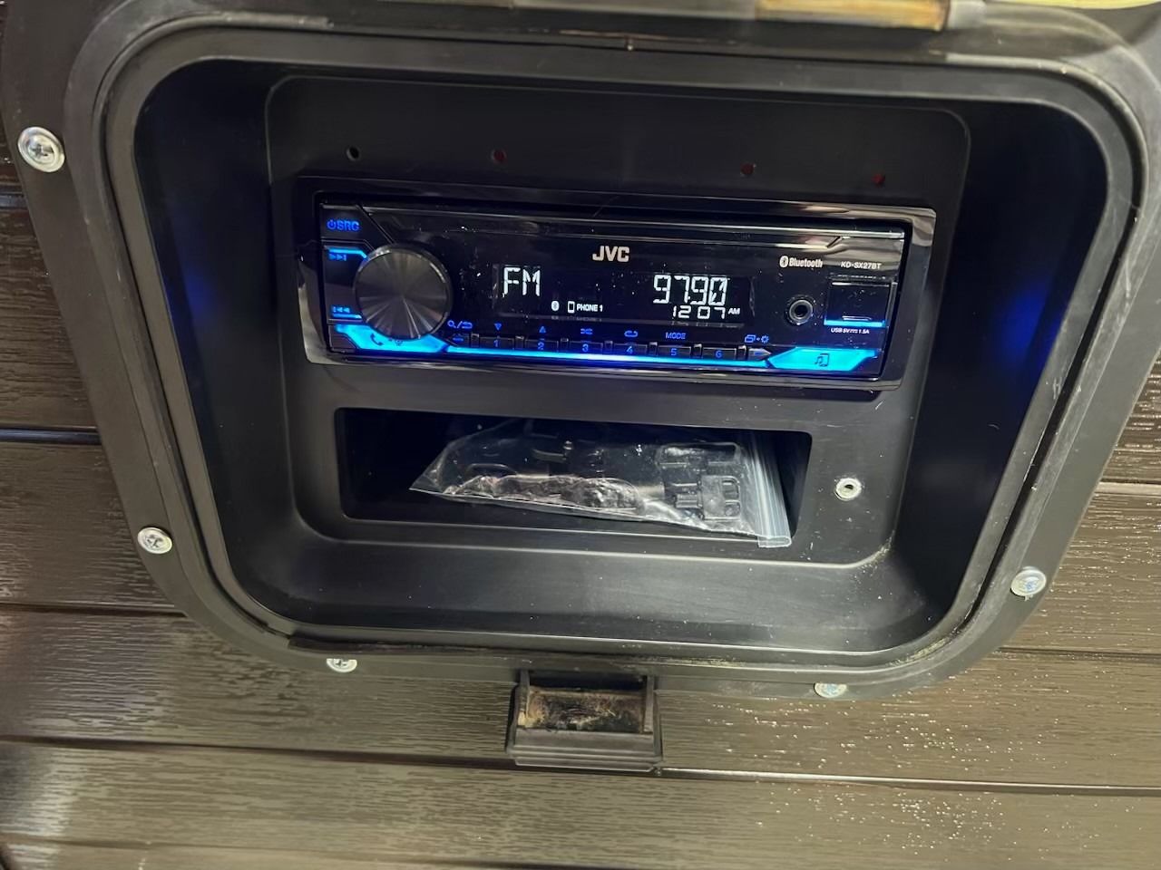JVC car stereo installed in a black compartment, displaying FM 97.9, with a small storage area below.