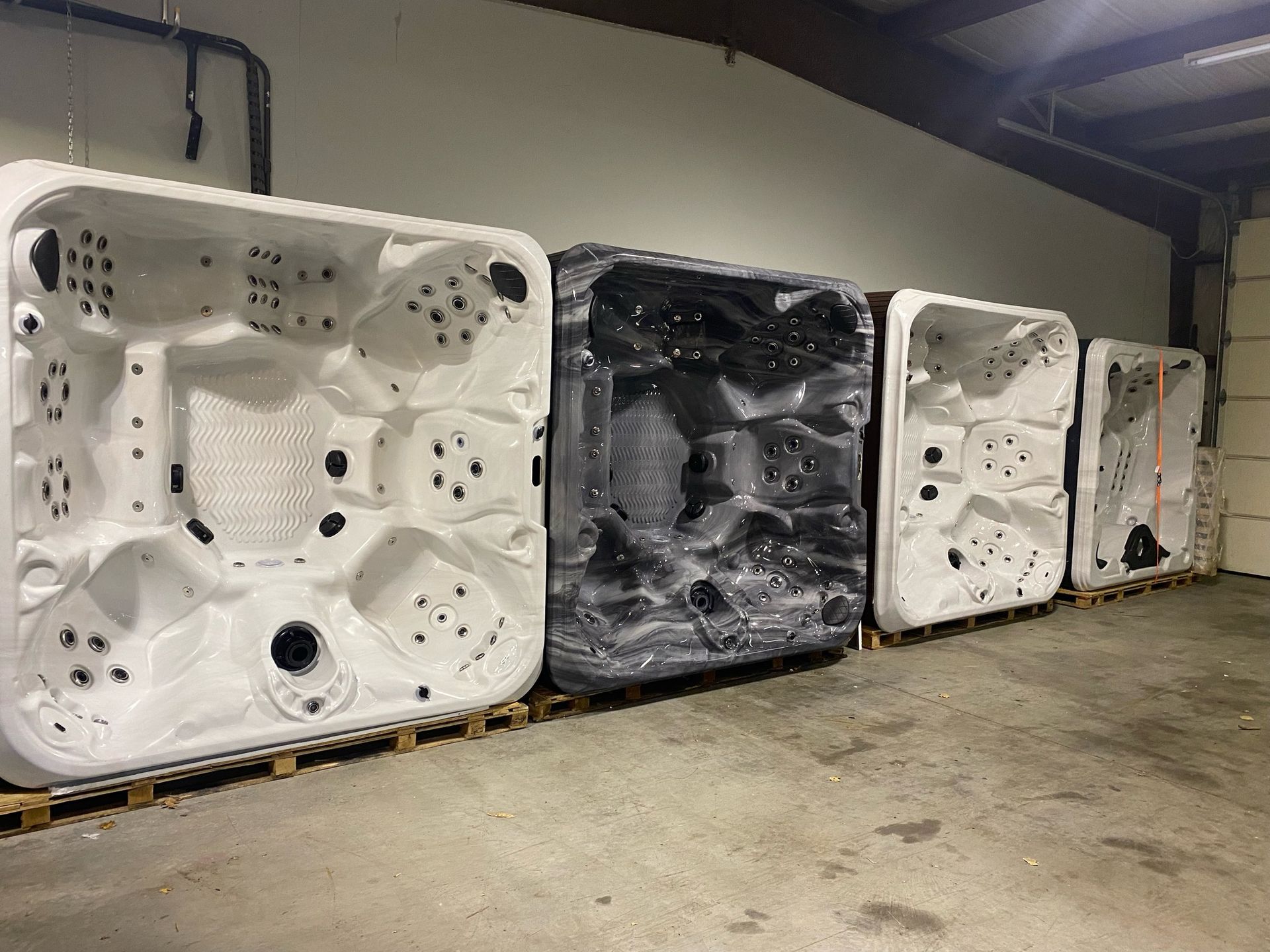 Four hot tubs in a row, two white, one black, and one covered, indoors on pallets.