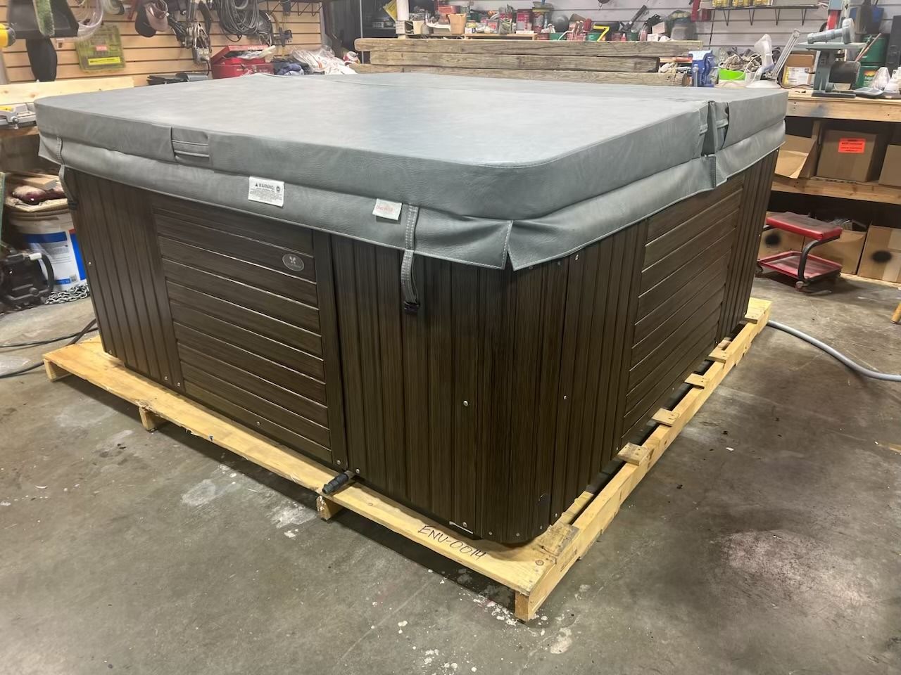 Hot tub with gray cover on a wooden pallet in a workshop setting. Dark brown wooden siding.