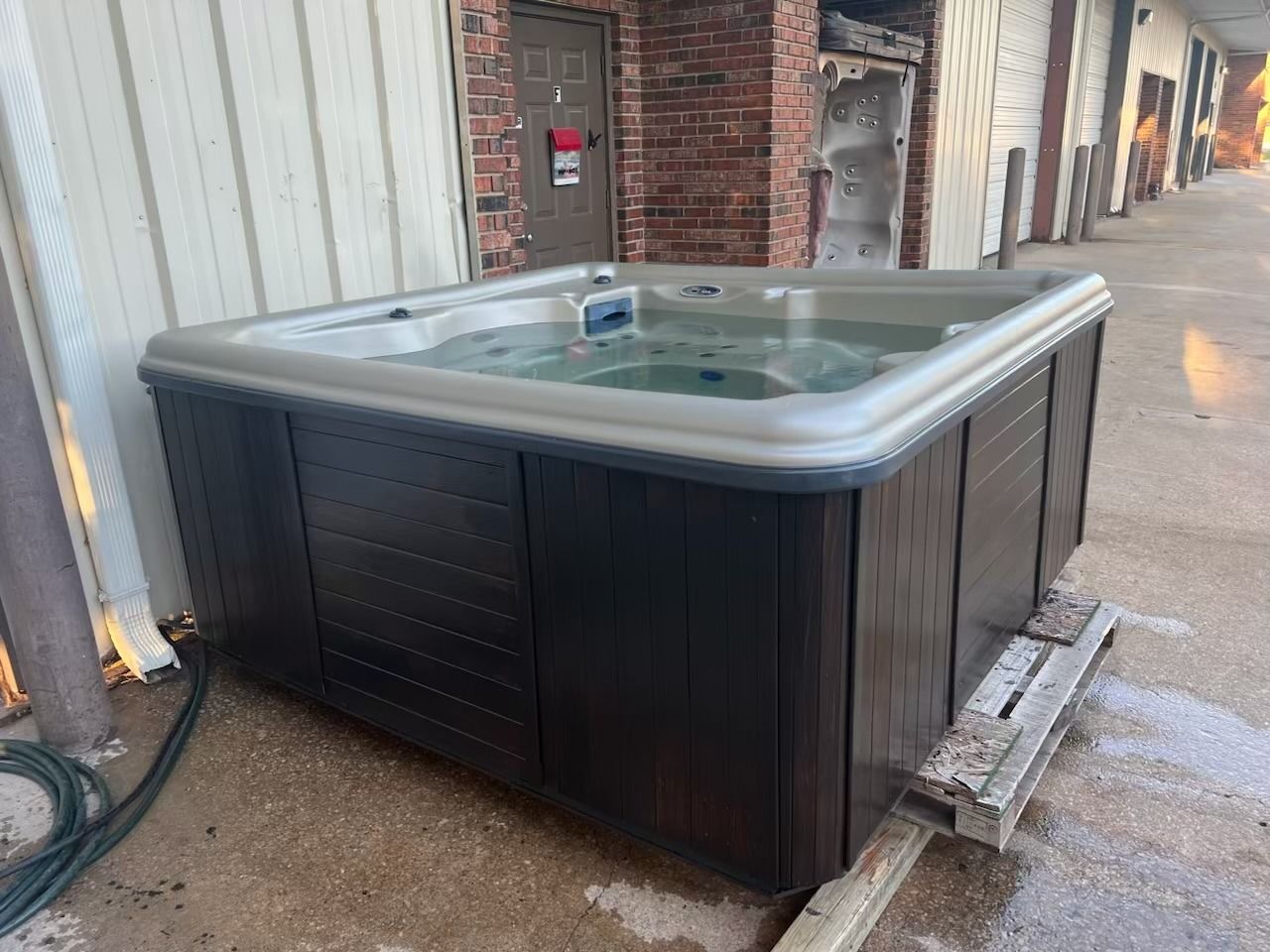 A brown hot tub filled with water sits outdoors next to a brick building.