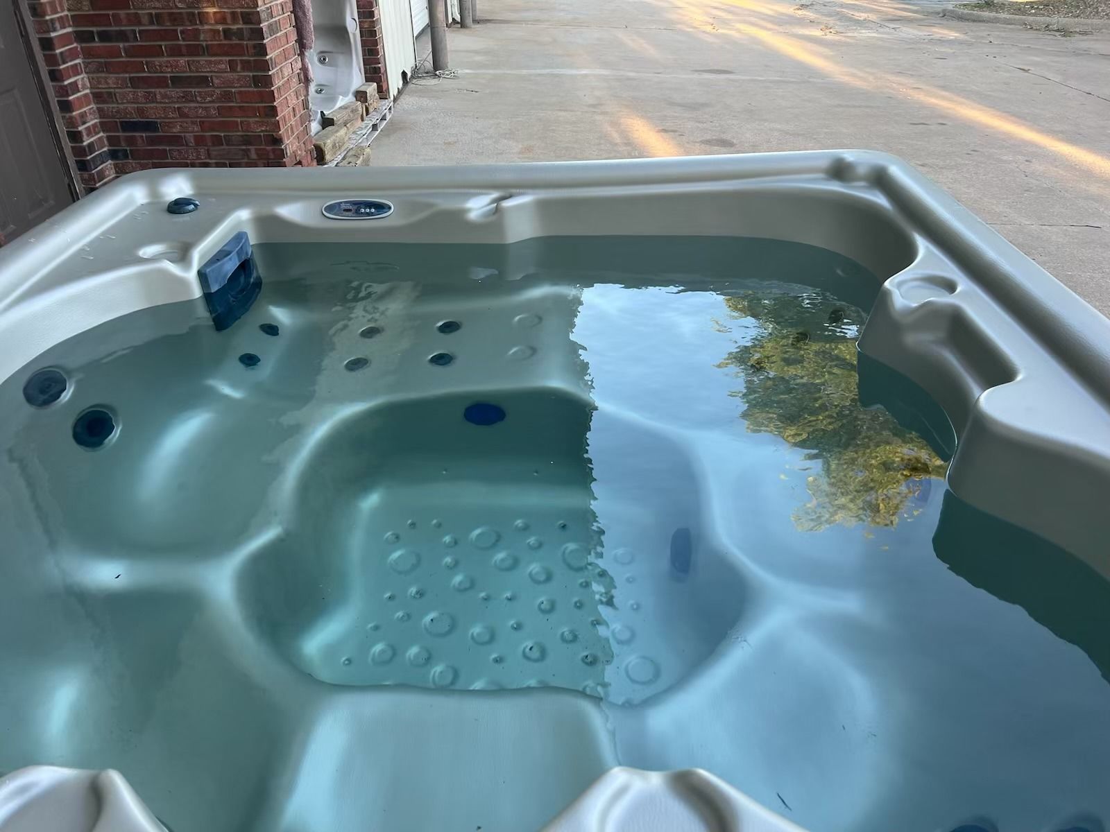 A light beige hot tub filled with water, outdoors near a building, water reflecting trees.