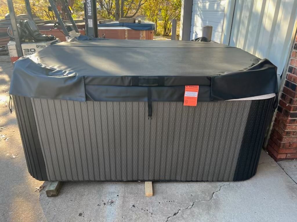 Dark brown hot tub with black cover, outdoors, on wooden beams, next to a brick wall.