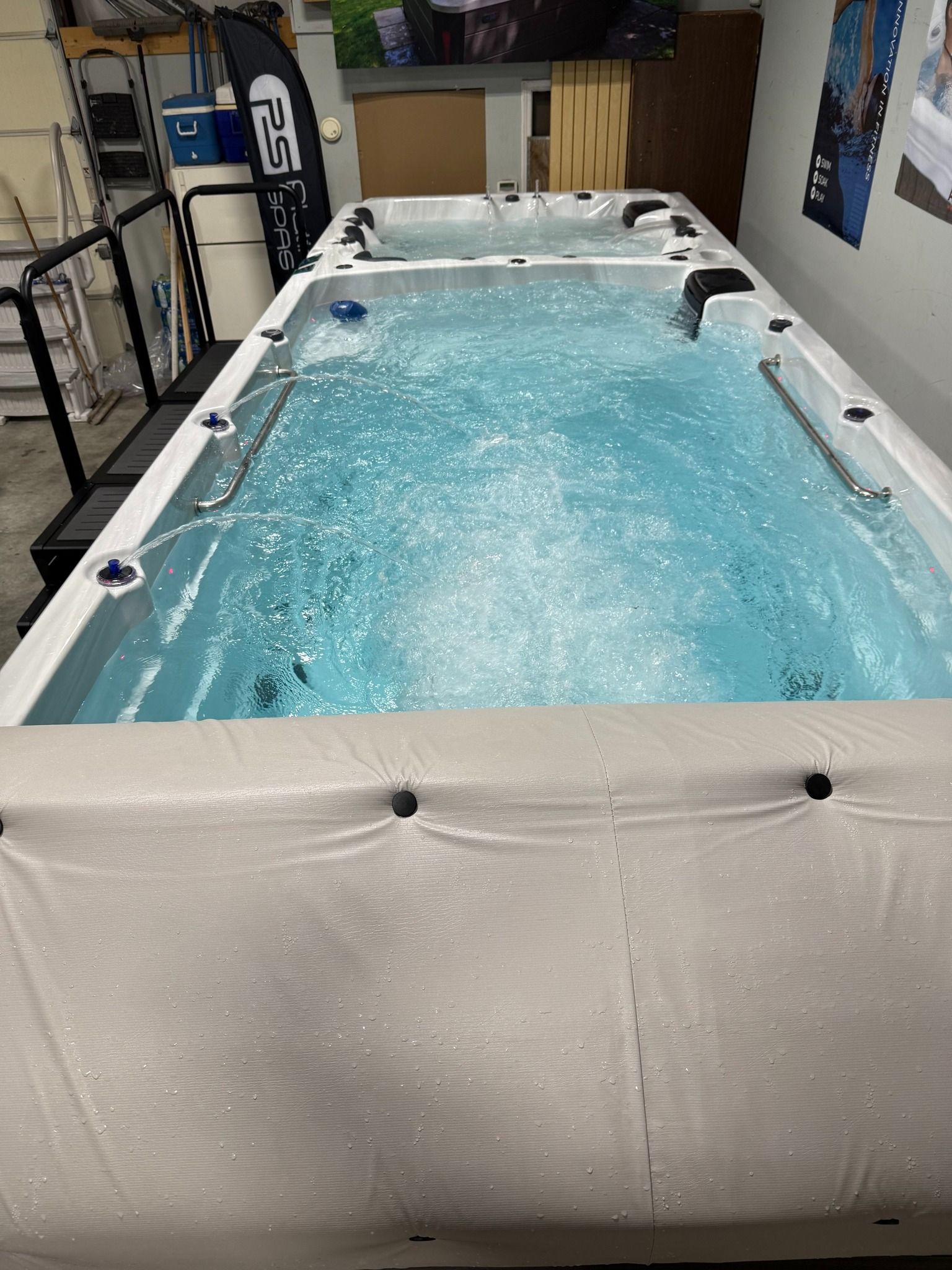 A long, rectangular, indoor swim spa filled with bubbly turquoise water; a white cover is on the side.