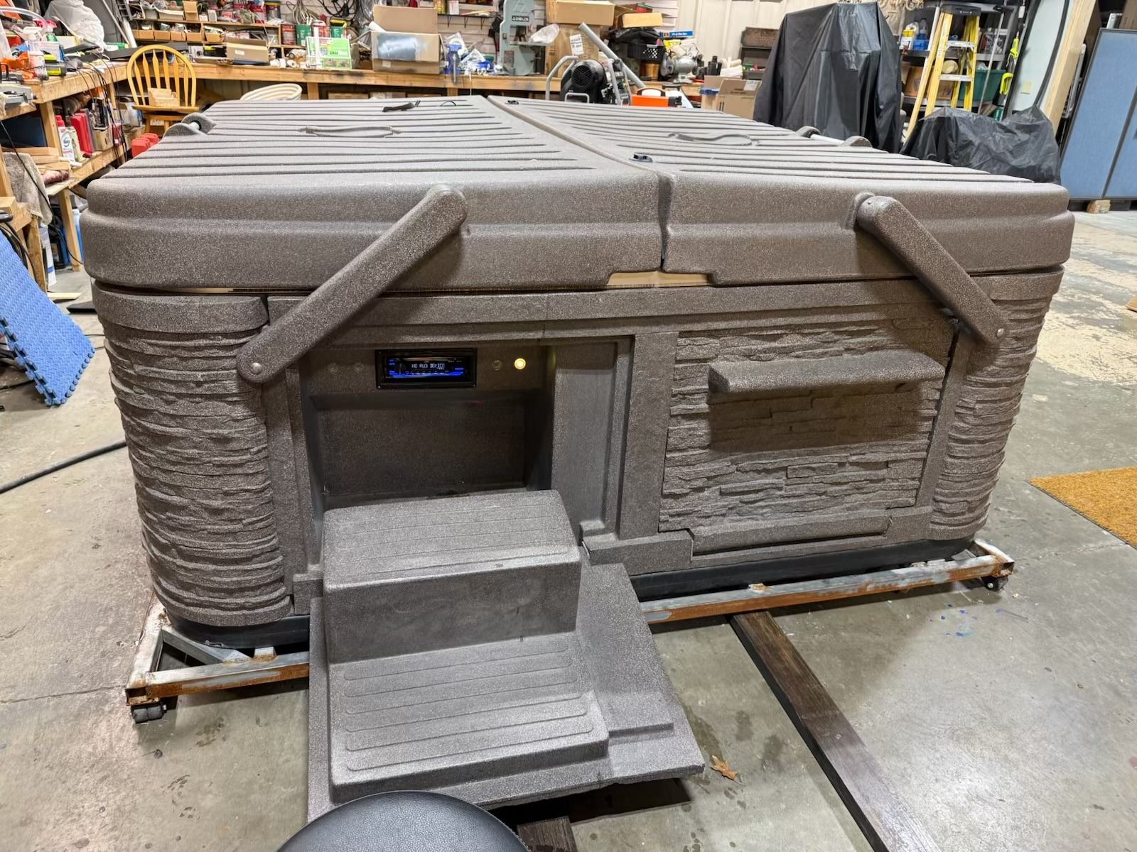 A brown, rectangular hot tub with steps and a control panel, inside a workshop.