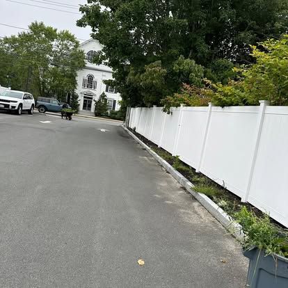 Quiet residential street with parked cars, white fence, and a white house in the background