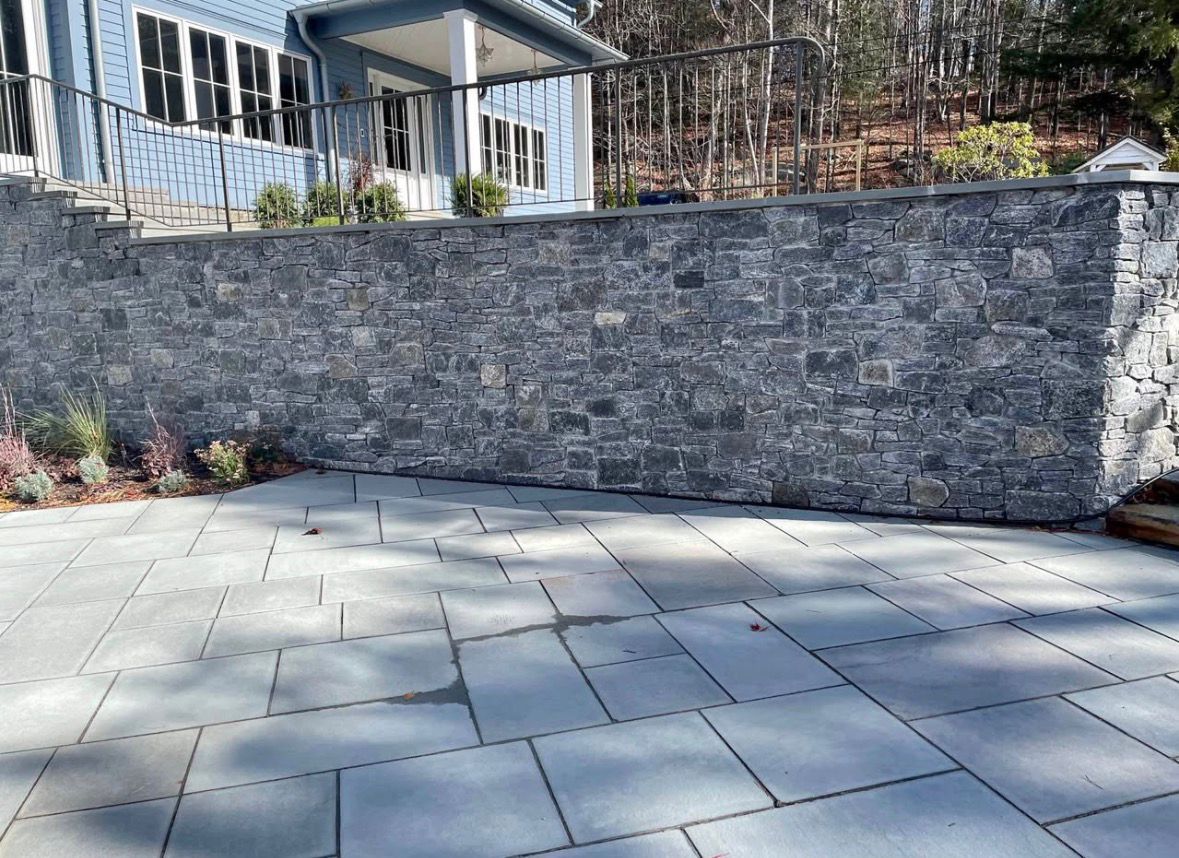 Stone retaining wall beside a paved patio in a residential backyard with a house and deck in the background