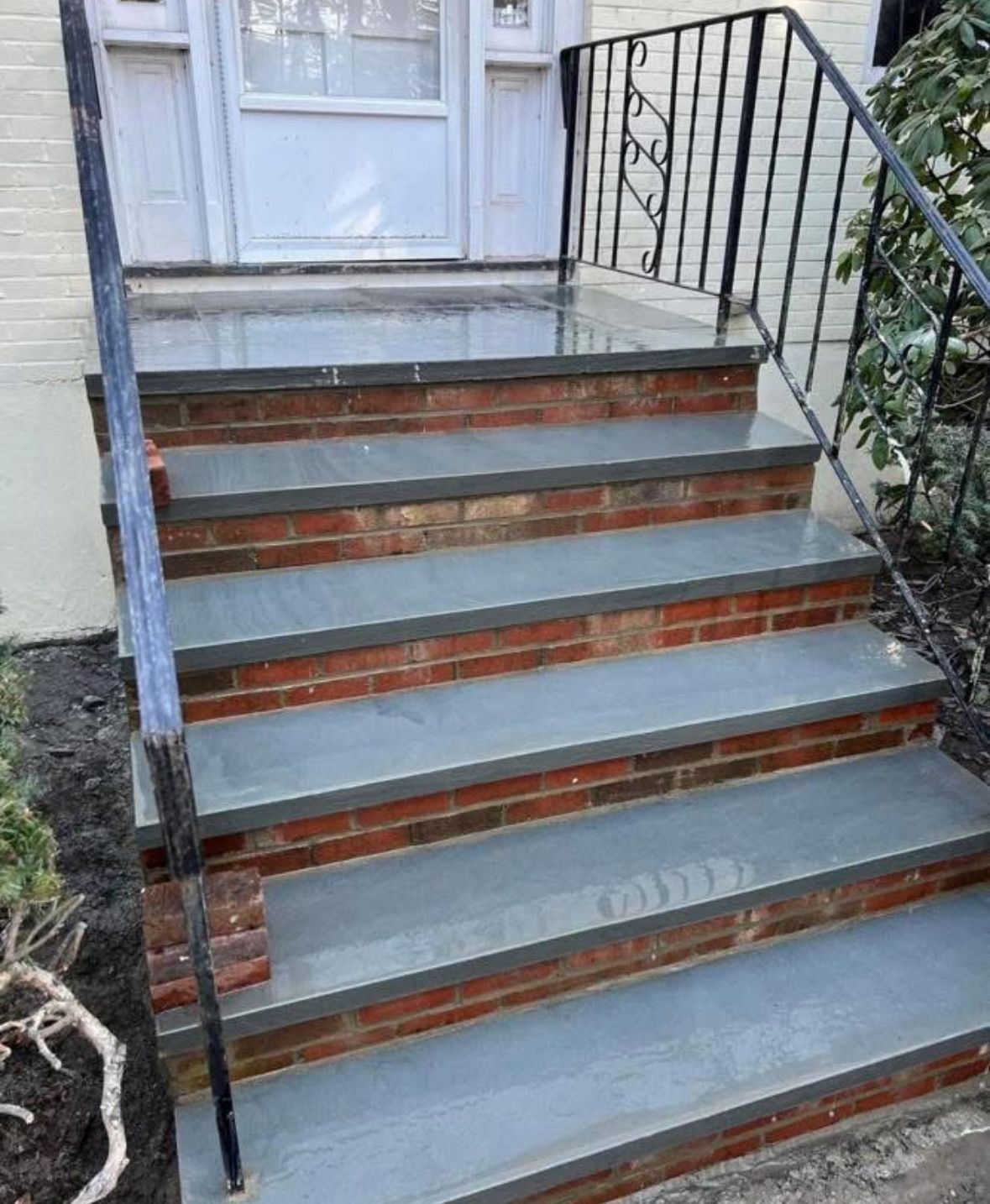 Front steps with gray treads, brick risers, and black railings leading to a white front door.