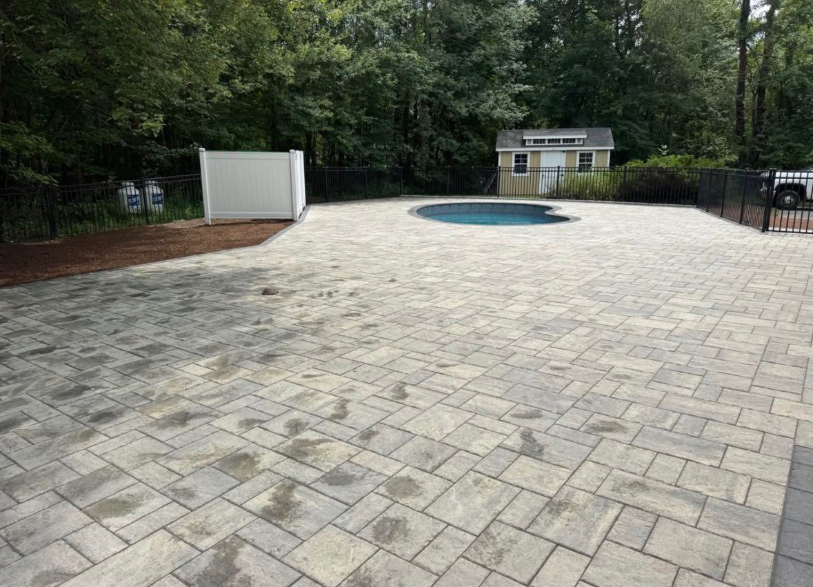 Paved backyard patio with a circular pool or hot tub, bordered by trees and a small shed.
