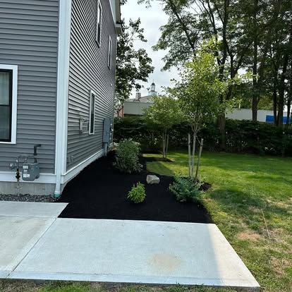 Side of gray house with concrete patio, black mulch landscaping bed, shrubs, and green lawn.