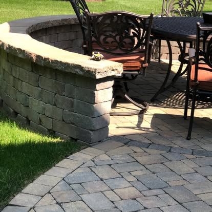 Curved stone patio wall beside black metal chairs on a sunlit brick patio and lawn