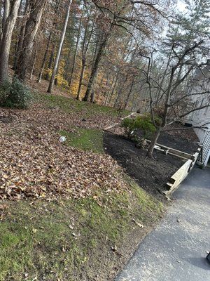 Leaf-covered wooded hillside beside a paved path and a small dark-roofed structure with stairs