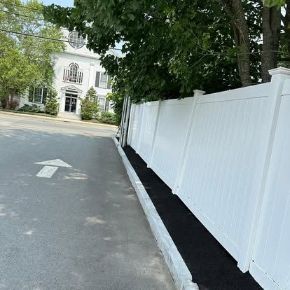Tree-lined street beside a white fence and sidewalk, with a white building ahead.