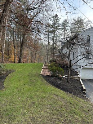 Sloped green lawn beside a white house and driveway, with leafless trees in the background.