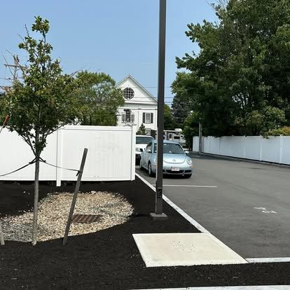 Parking lot with a light blue car, white fence, and small trees beside a road.
