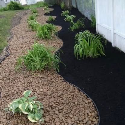 Curved rock mulch border separating green plants from black landscape fabric along a white fence.
