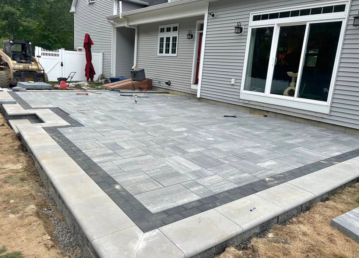 Backyard stone patio under construction beside a house, with large gray pavers and a concrete border