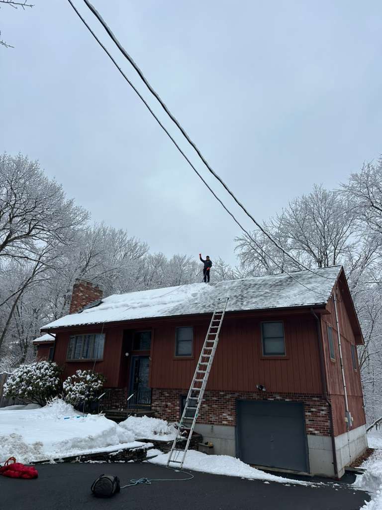 Person on a snowy roof beside a ladder on a red house with a garage, under power lines
