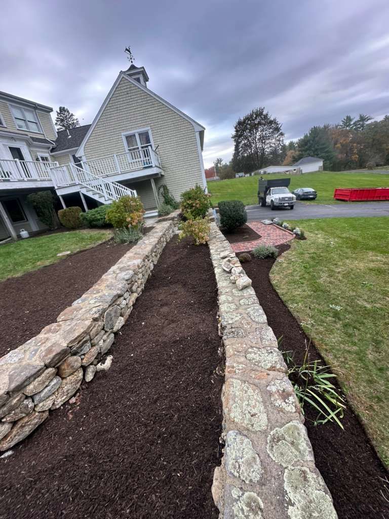 Stone-bordered garden beds beside a gray house, with mulch, lawn, and a driveway in the background.