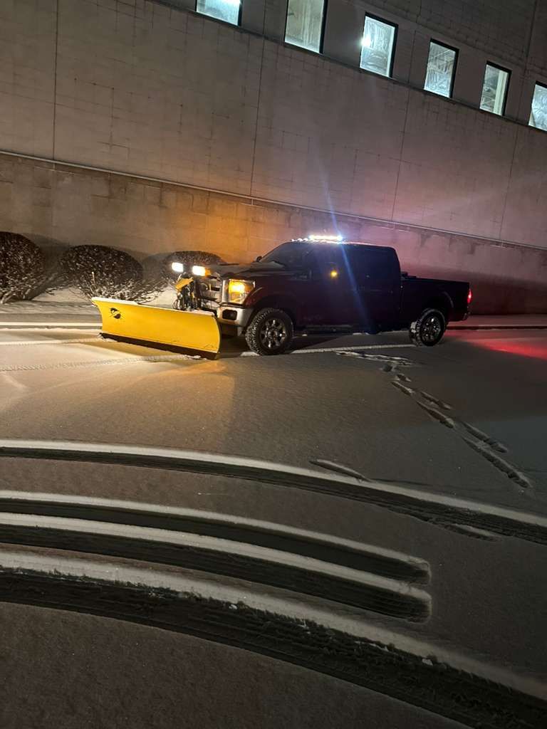 Black pickup with a yellow snowplow blade parked on a road at night by a concrete wall