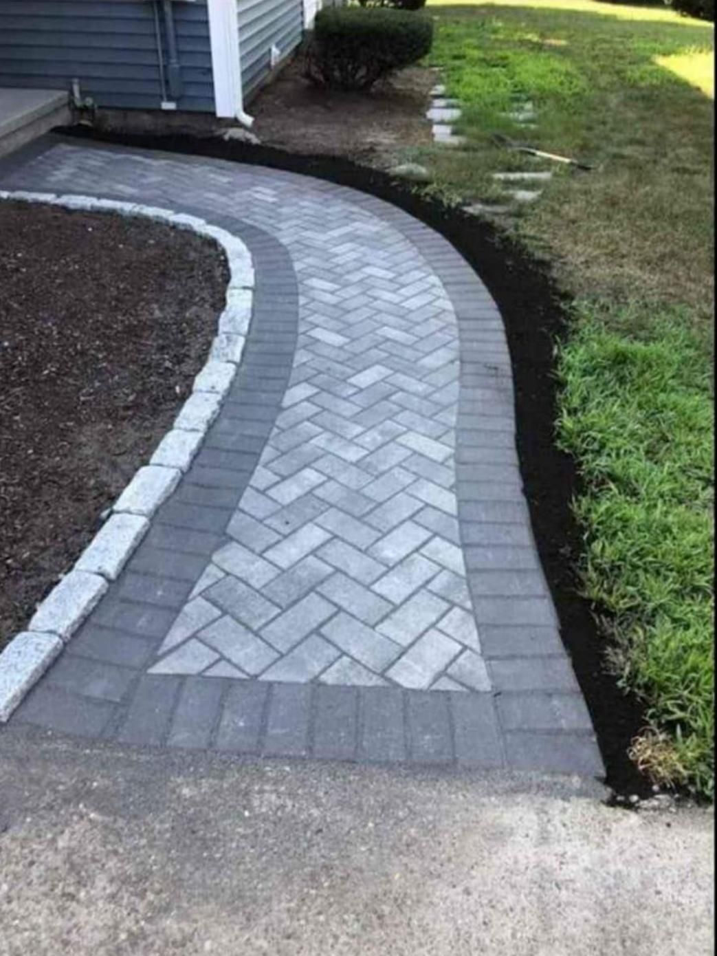 Curved gray brick walkway beside a house and lawn, bordered by dark edging.