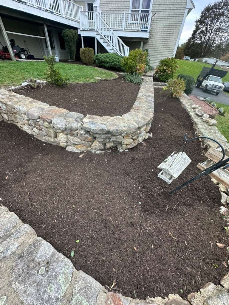 Stone-edged front garden bed with dark mulch, small plants, and a house porch in the background