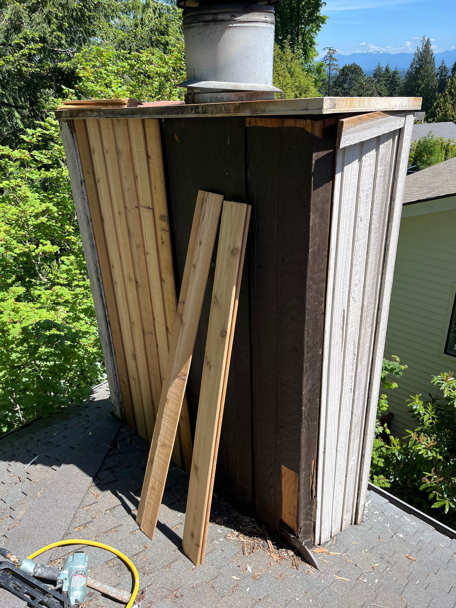Wooden fence panels leaning against a shed beside a driveway, with a vent pipe on top and a hose nearby.