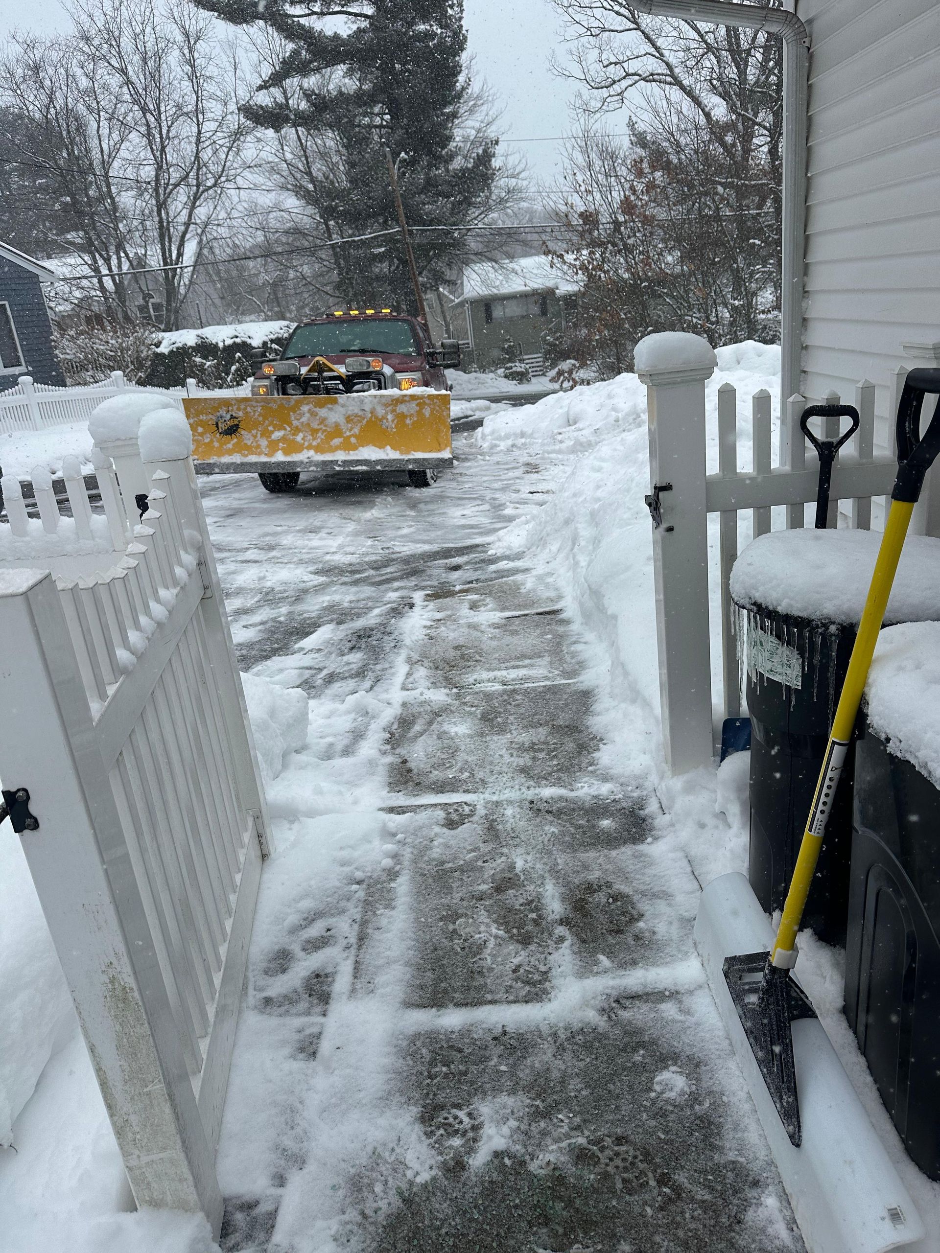Snowy front walkway with a yellow truck parked in the driveway, seen through an open white gate.
