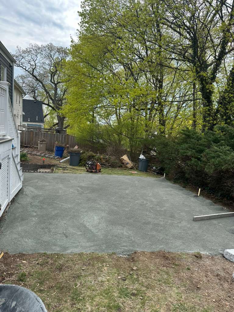 Backyard with a paved patio, grass edges, and tall green trees beside a house