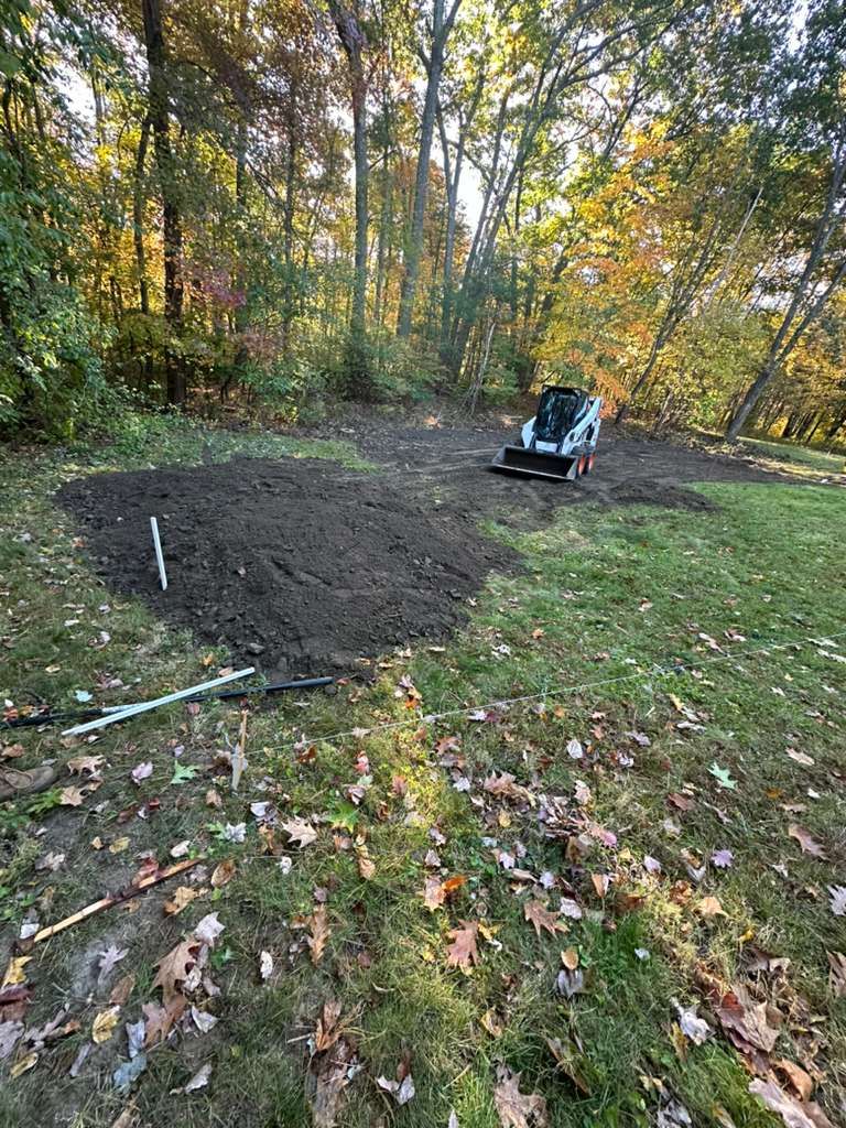 Freshly tilled soil patch in a wooded yard, with a small black garden machine nearby.