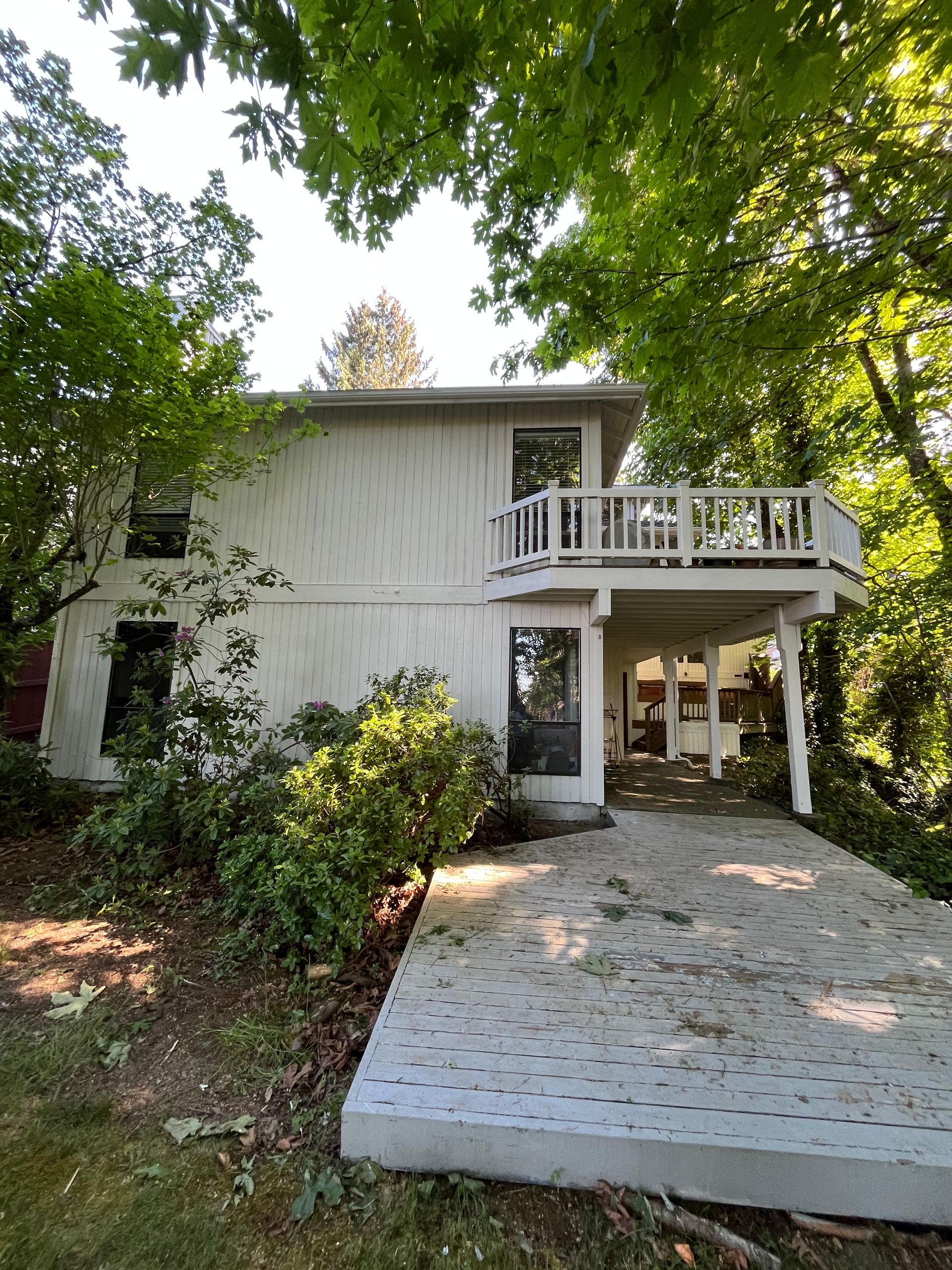 Two-story house with white siding, balcony, and driveway surrounded by trees and greenery