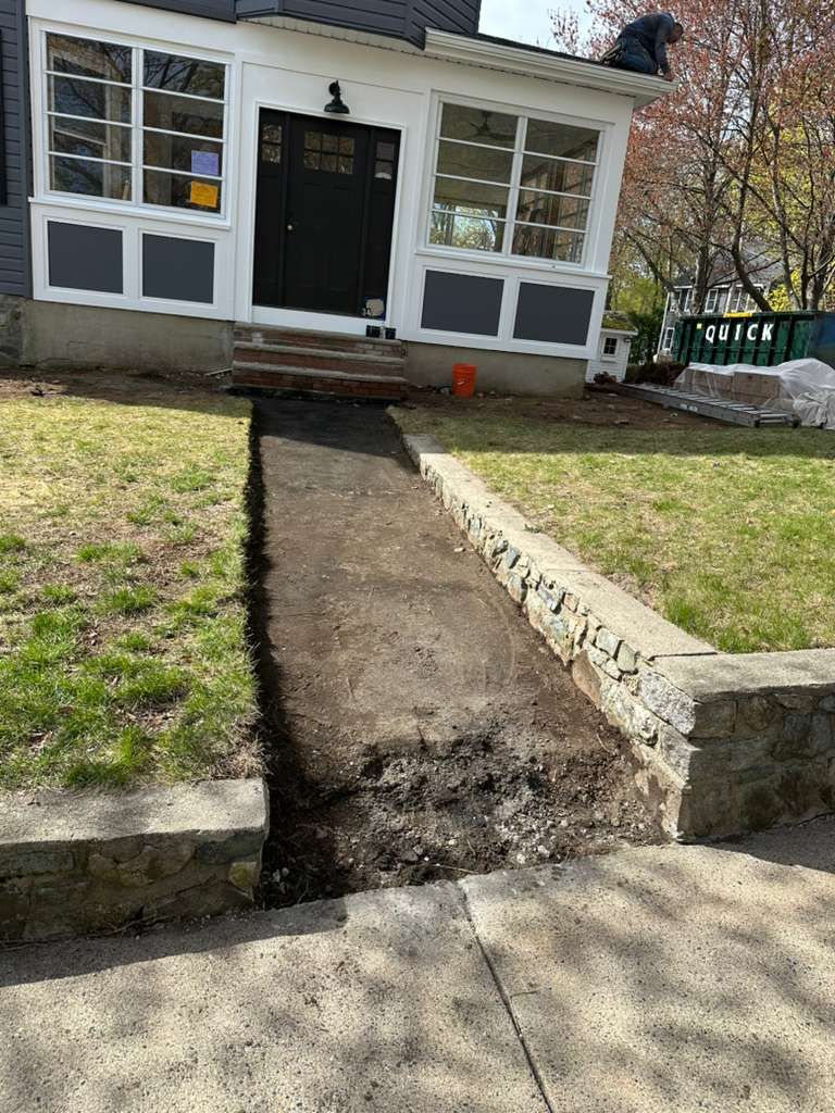 Front yard with a newly dug dirt trench leading to a house entrance beside a small stone retaining wall.
