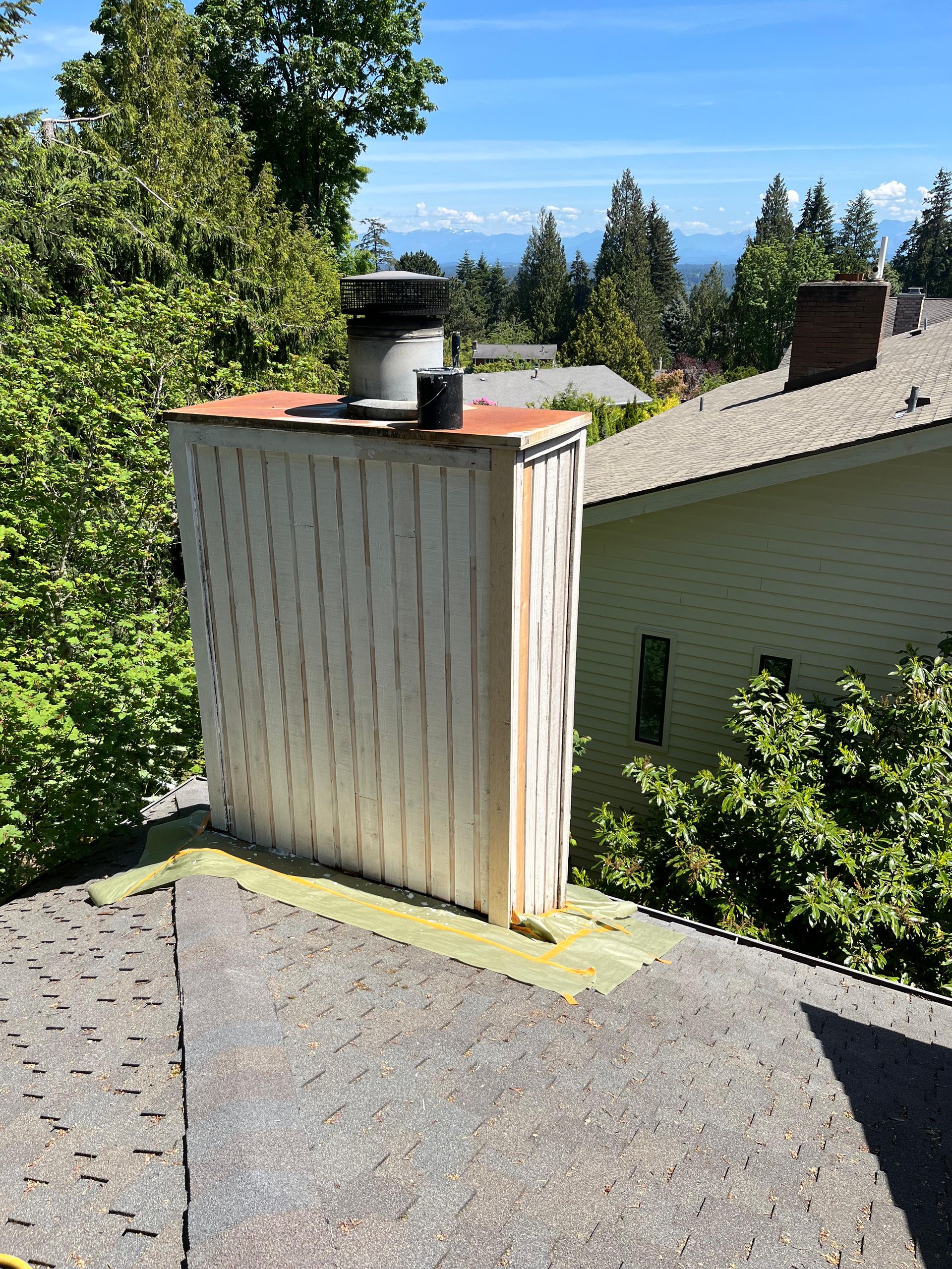 White rooftop chimney vent beside a green house, with trees and blue sky in the background
