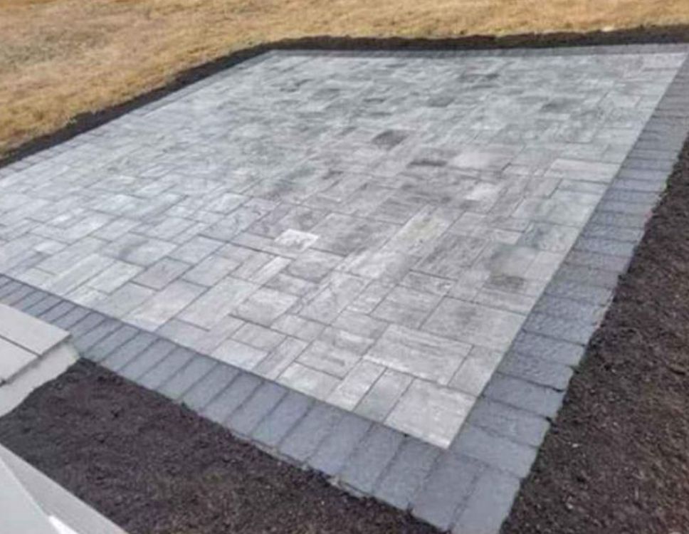 Rectangular gray stone patio bordered by dark pavers and surrounded by soil