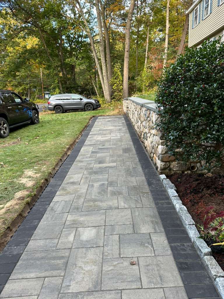 Stone walkway beside a hedge and retaining wall, with parked cars and trees along a grassy yard