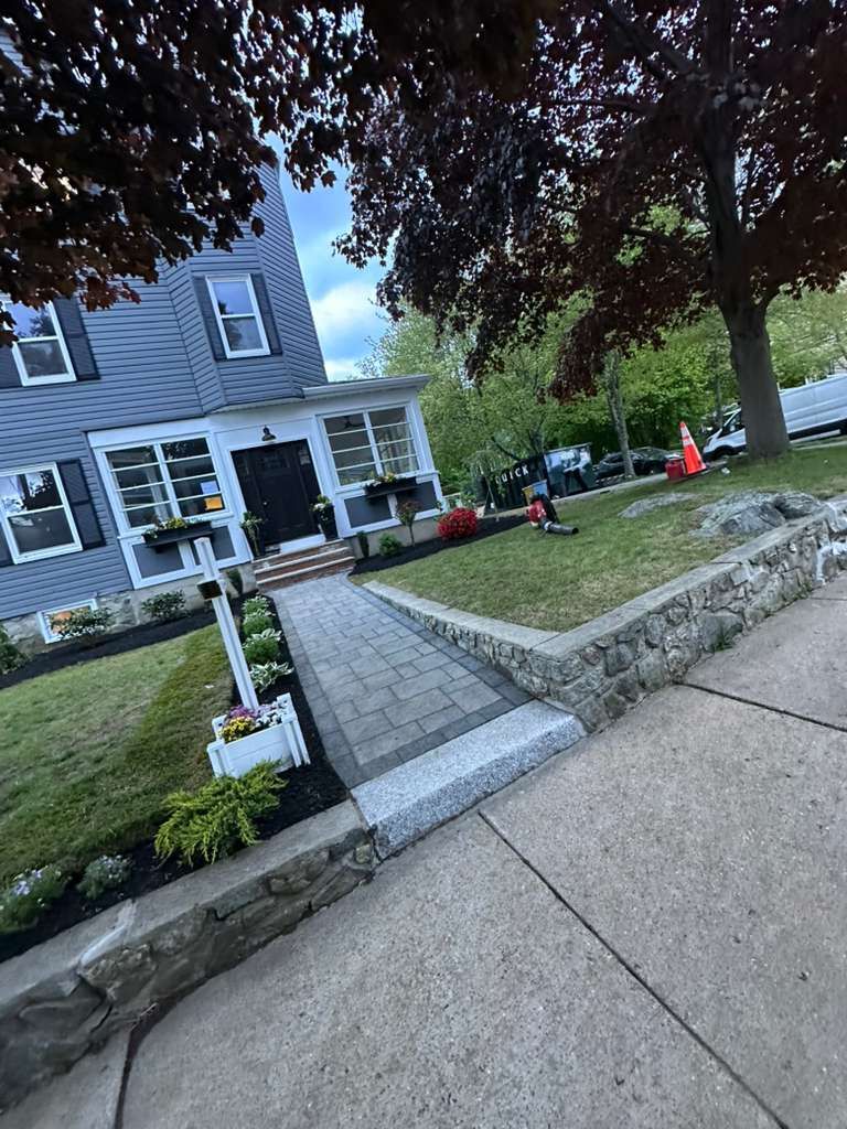Sidewalk leading to a gray house with flower beds, a sloped lawn, and orange cones near the entrance.