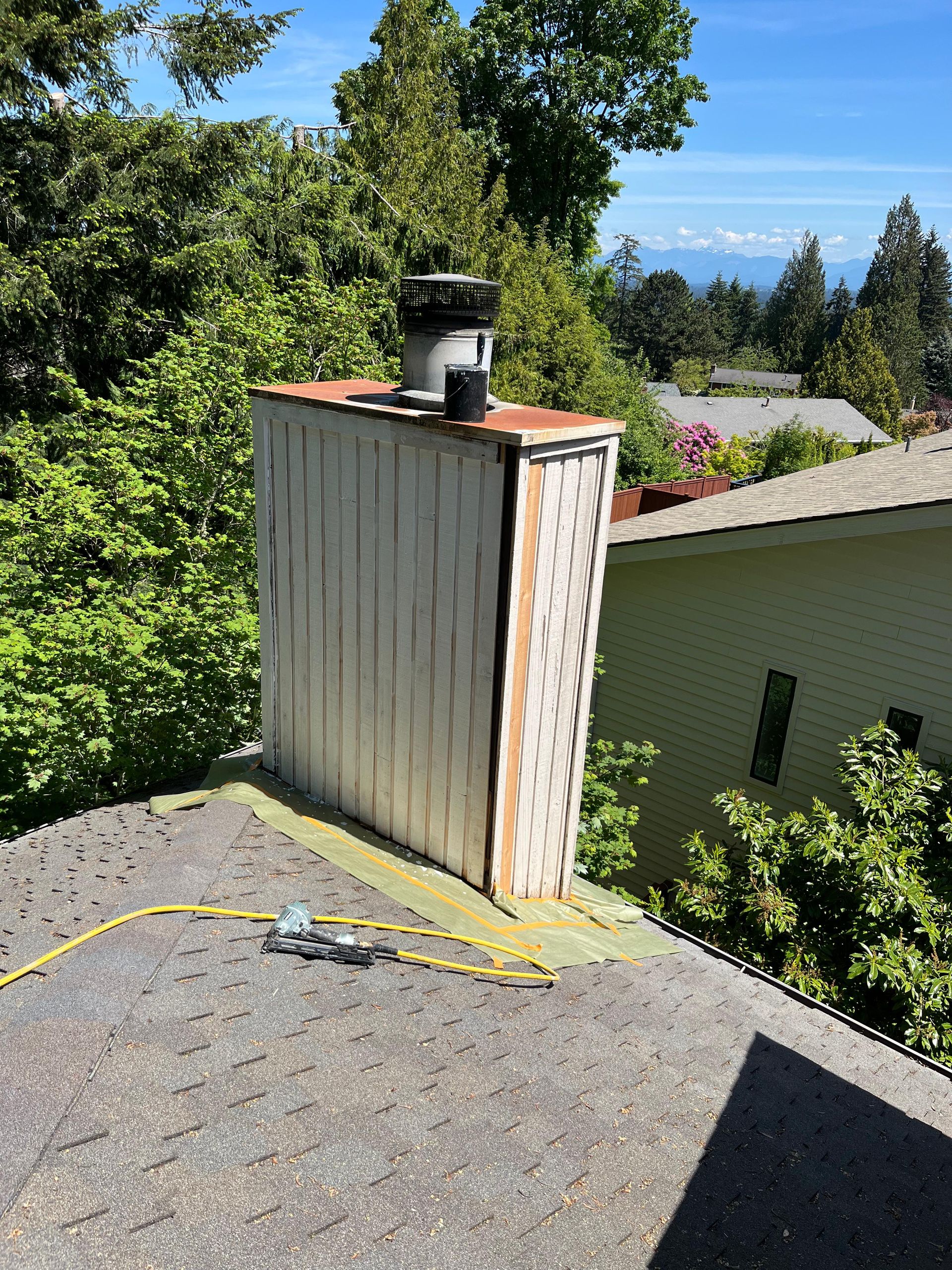 Rooftop vent flue on a gravel roof beside trees, with a yellow hose and ocean view in the distance