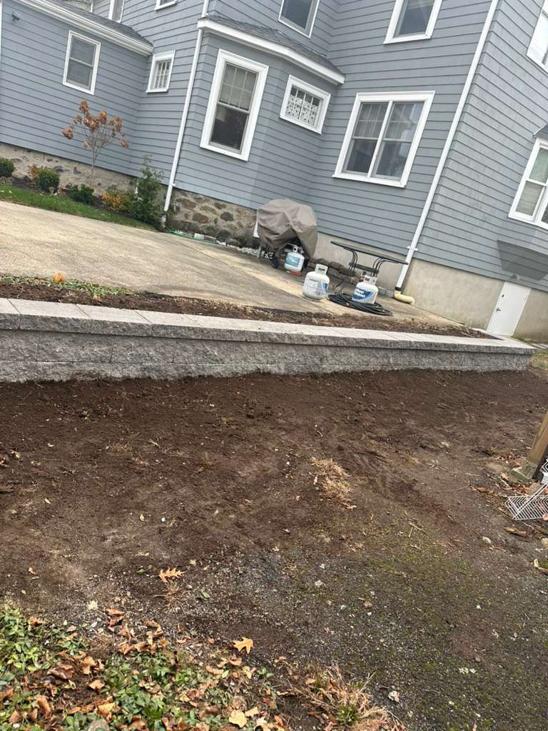 Freshly graded dirt lot beside a gray house with patio chairs and a concrete walkway