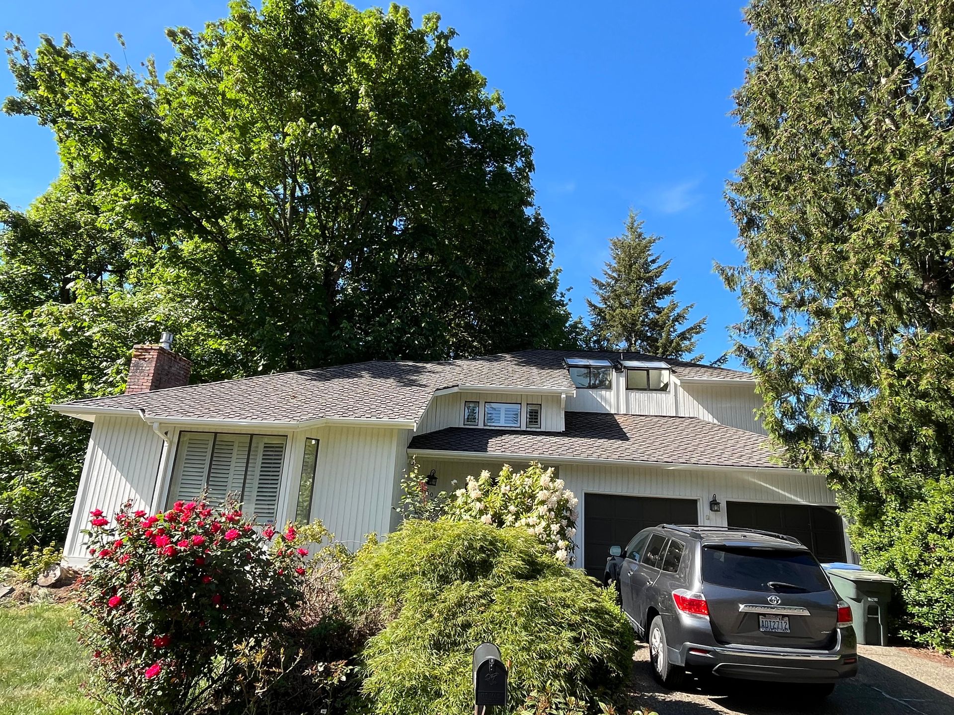 White house with a gray SUV in the driveway, surrounded by lush trees and pink flowers under a blue sky