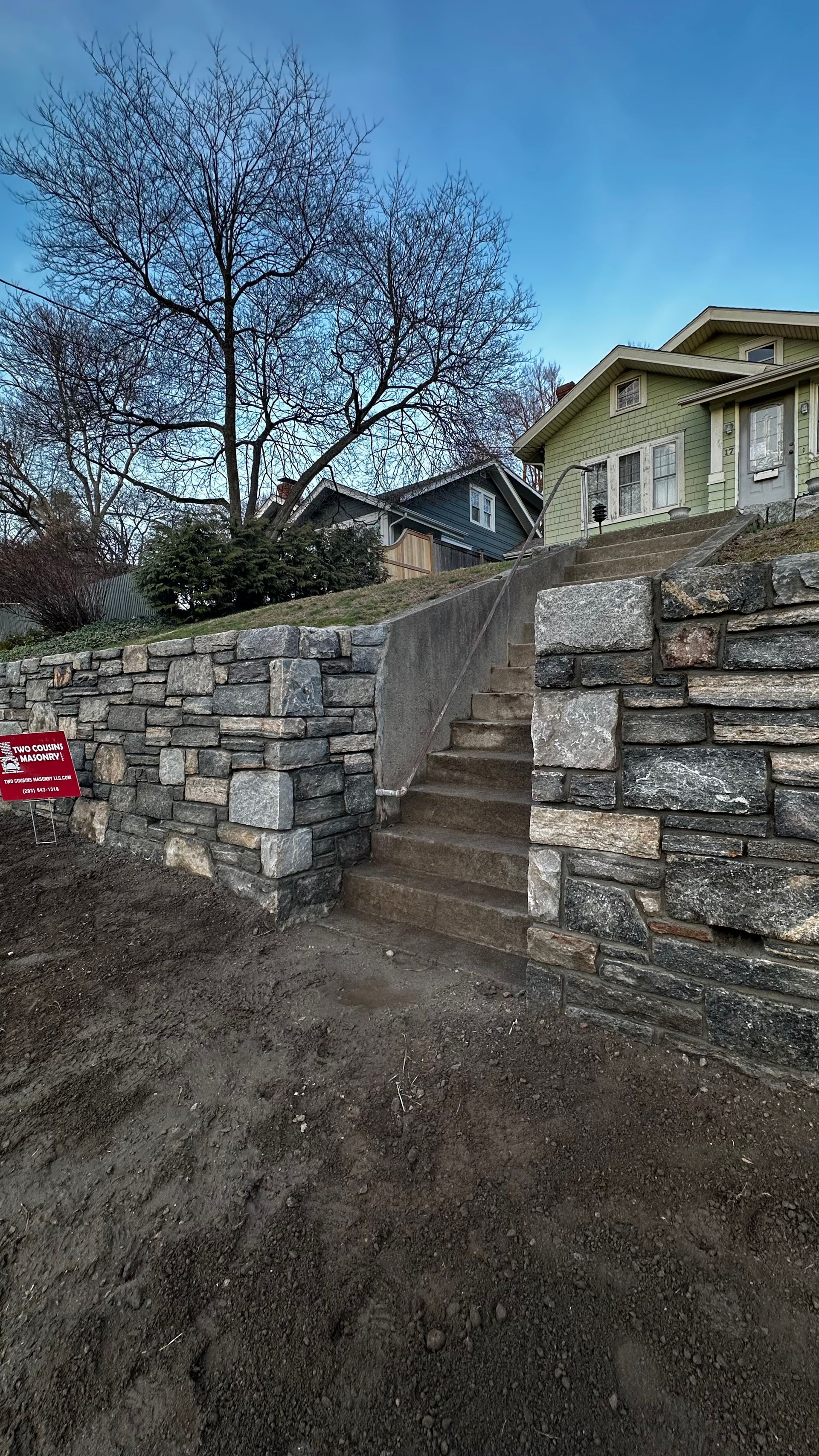 Stone stairway between retaining walls leading to houses, with a leafless tree at dusk.