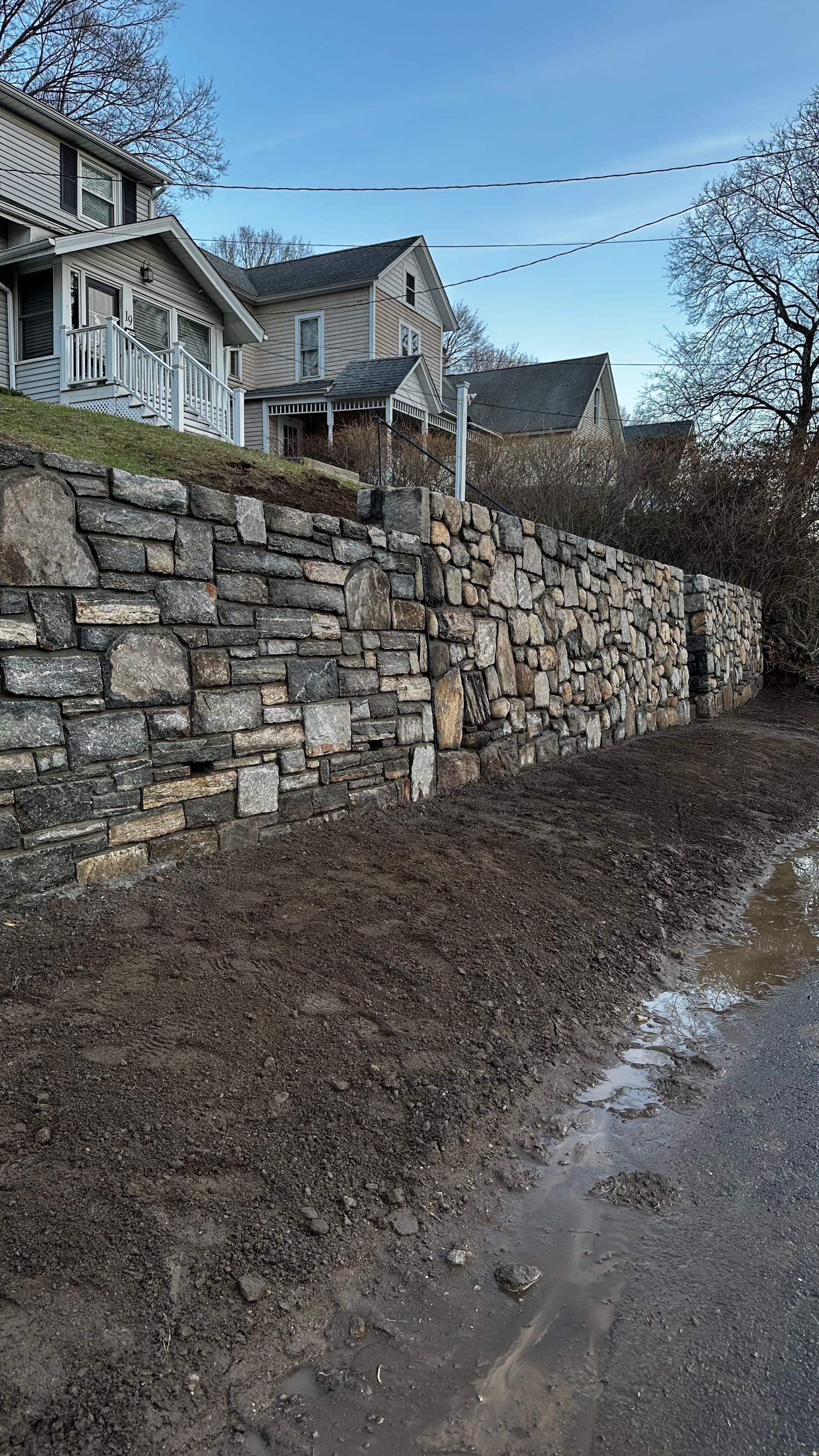 Stone retaining wall beside a muddy path, with houses on a hillside under a blue sky.