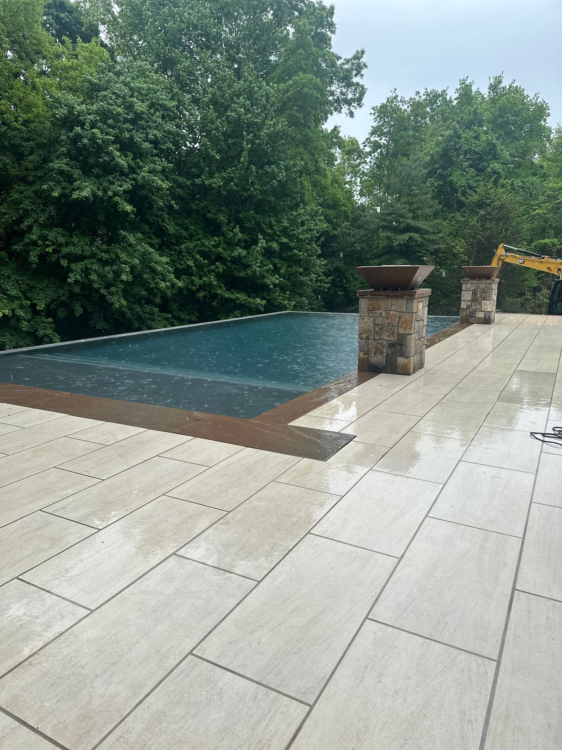Outdoor patio with stone pavers, a covered pool, and brick pillars beside dense green trees
