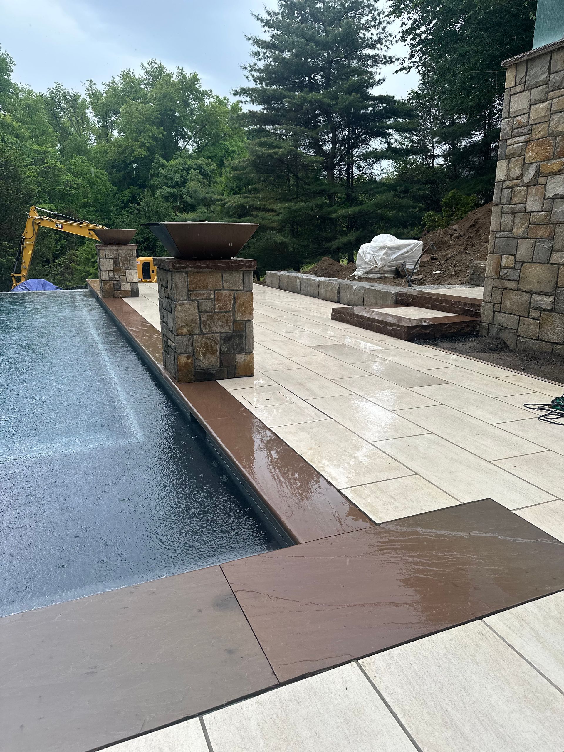 Backyard patio with wet stone pavers, pool edge, stone pillars, and trees beyond