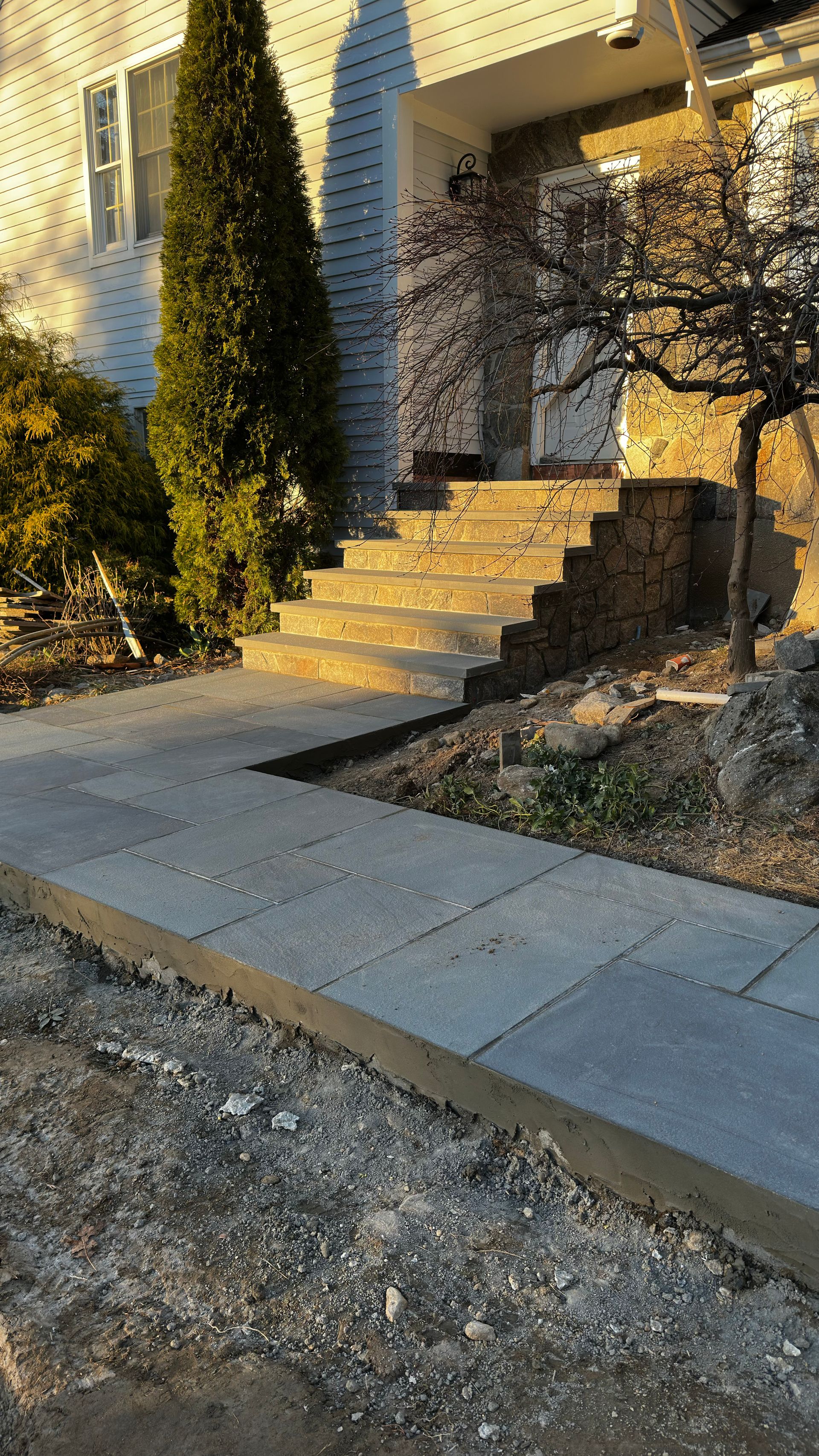 Front steps with stone railing beside a house, lit by warm sunlight, with a sidewalk and gravel foreground.